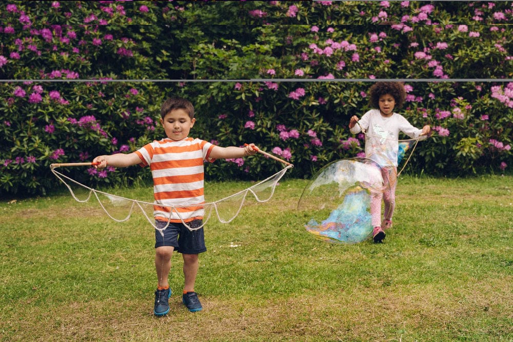 two children are playing in a garden on the grass, they are each holding a bubble wand, one girl has a large bubble that is half her size.