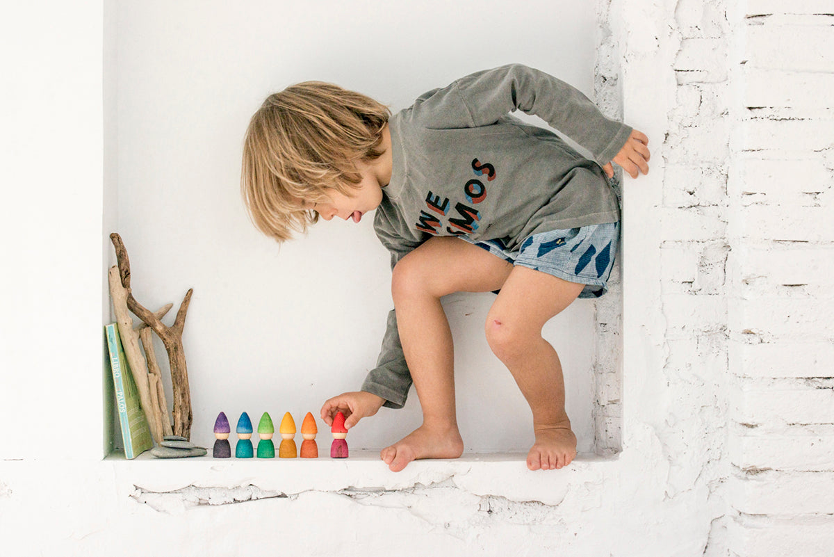 A small child engrossed in play. He is leaning over towards 6 small wooden people in rainbow colours