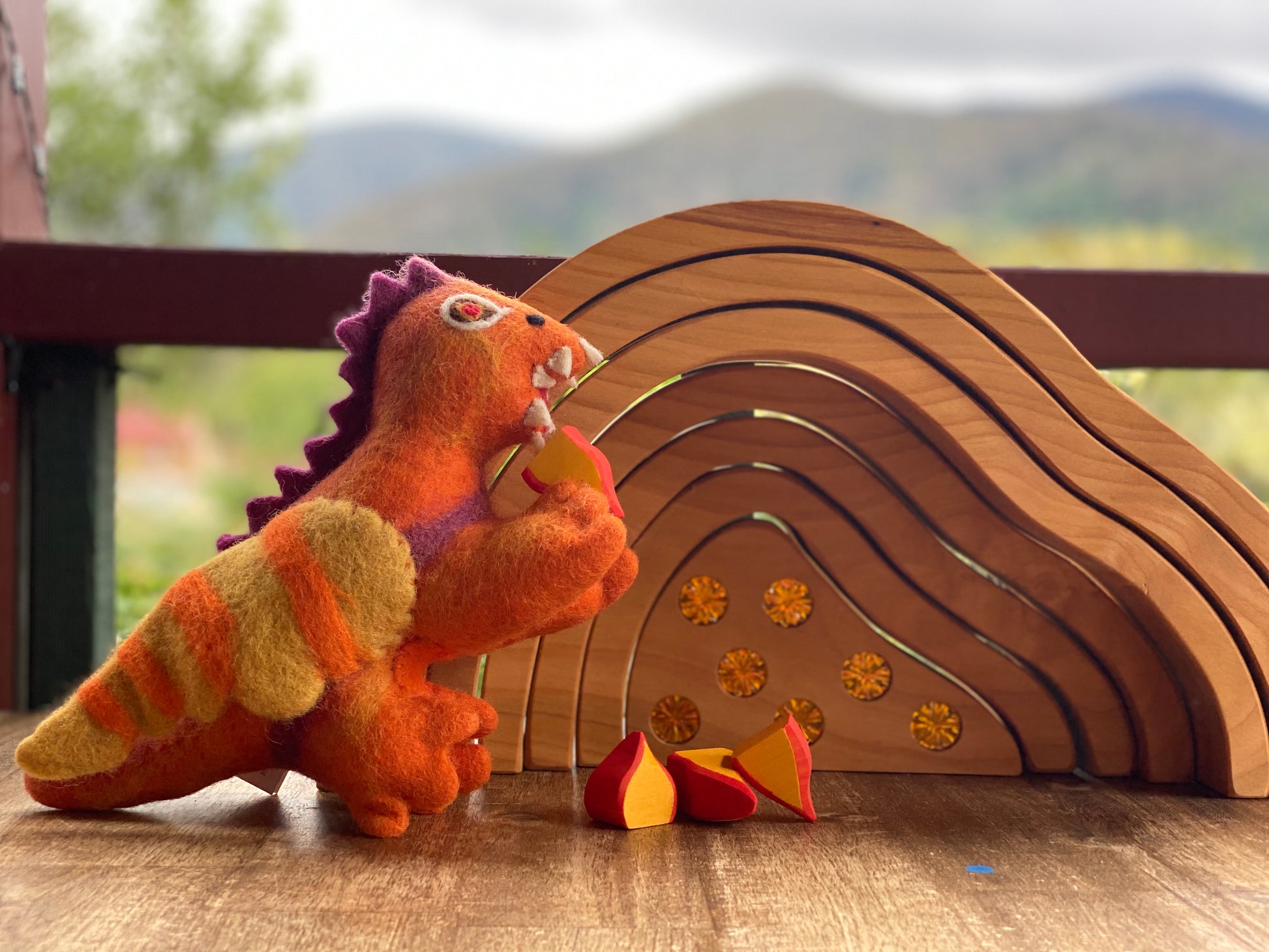 A handmade orange felt toy dragon warming itself by small wooden fire shaped pieces painted orange and yellow.  Behind the dragon is a handcrafted wooden cave made of 7 concentric pieces. The smallest piece is embellished with 7 orange sparkly gems.